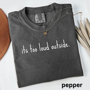 May include: A folded pepper-colored t-shirt with the text "its too loud outside" in white. A silver necklace with a pendant is on the shirt. The shirt has a Comfort Colors tag.