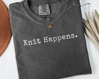 May include: A dark gray t-shirt with the phrase "Knit Happens" in white lettering. A silver necklace with a small pendant rests on the shirt. The word "pepper" is printed at the bottom.