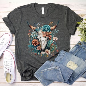 Puede incluir: Una camiseta gris oscuro con un dise&ntilde;o de calavera floral. La calavera est&aacute; rodeada de flores en tonos azules, rosas y naranjas. El dise&ntilde;o est&aacute; centrado en la camiseta.