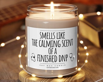 May include: A brown soy wax candle with a white label that reads "Smells like the calming scent of a finished DNP". The label also includes a stethoscope icon on each side. The candle is sitting on a wooden coaster.
