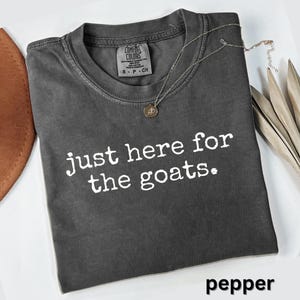 May include: A dark gray Comfort Colors t-shirt with the text "just here for the goats." A silver necklace with a pendant rests on the shirt. The word "pepper" is at the bottom.