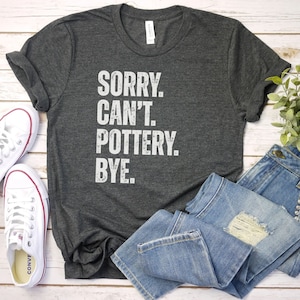 May include: A dark gray t-shirt with the text "SORRY. CAN'T. POTTERY. BYE." printed in white.