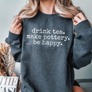 May include: A dark gray sweatshirt with the text "drink tea. make pottery. be happy." printed in white.