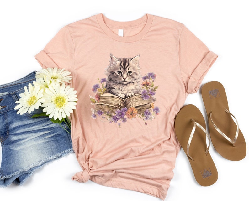 May include: A pink t-shirt with a graphic of a cat reading a book surrounded by flowers. The cat is gray and white with green eyes. The book is brown and has a white cover. The flowers are purple, pink, and yellow.