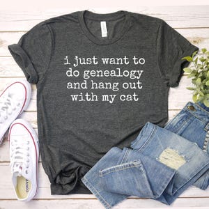 May include: A dark gray t-shirt with the text "i just want to do genealogy and hang out with my cat" printed in white.