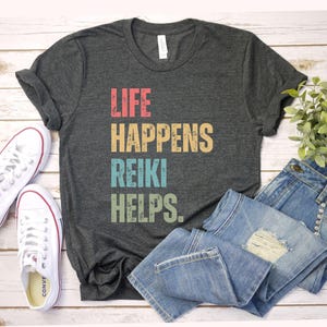 May include: Dark gray t-shirt with the text "LIFE HAPPENS REIKI HELPS" in colorful, distressed lettering. The shirt is paired with blue jeans and white canvas sneakers, creating a casual, everyday look.