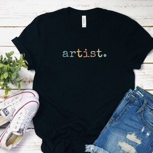 Peut inclure: T-shirt noire avec le mot "artiste" imprim&eacute; en couleurs arc-en-ciel sur le devant. Le t-shirt est pos&eacute; &agrave; plat sur une surface en bois blanc avec une paire de baskets blanches et un short en jean bleu.