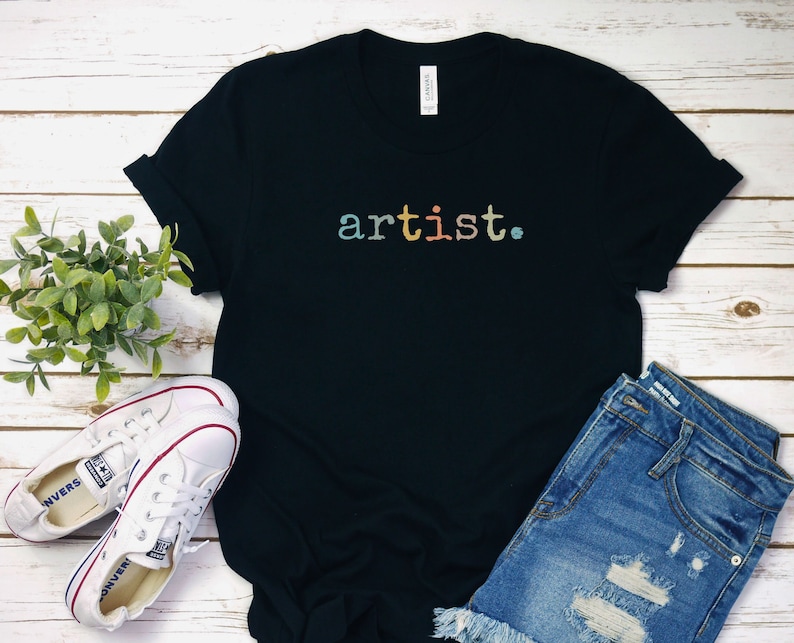 Peut inclure: T-shirt noire avec le mot "artiste" imprim&eacute; en couleurs arc-en-ciel sur le devant. Le t-shirt est pos&eacute; &agrave; plat sur une surface en bois blanc avec une paire de baskets blanches et un short en jean bleu.