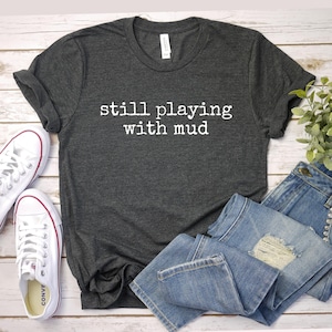 May include: Dark gray t-shirt with the text "still playing with mud" in white. The shirt is paired with light blue denim jeans and white canvas sneakers. The items are arranged on a light wood-grain surface.