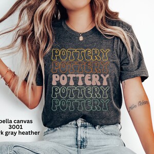 May include: A dark gray heather Bella Canvas 3001 t-shirt with the word "POTTERY" repeated five times in a rainbow of colors. The text is in a retro, bubble-like font.