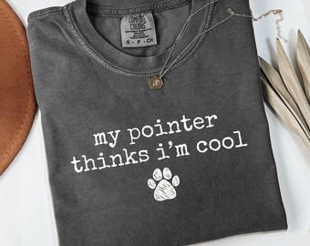May include: A dark gray Comfort Colors t-shirt with the text "my pointer thinks I'm cool" and a paw print graphic. A silver necklace with a pendant is draped across the shirt. The word "pepper" is at the bottom.