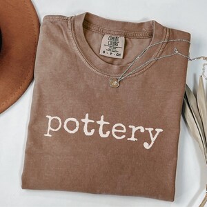 May include: A brown Comfort Colors t-shirt with the word "pottery" printed in white. A silver necklace with a small pendant is draped across the shirt. A brown hat is visible in the upper left corner.