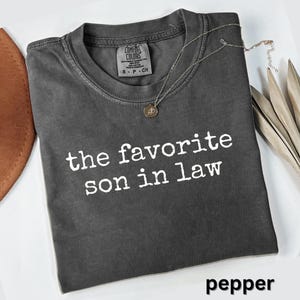 May include: A dark gray t-shirt with the text "the favorite son in law" in white. The shirt has a crew neck and a Comfort Colors tag. A silver necklace with a pendant rests on the shirt. The word "pepper" is at the bottom.