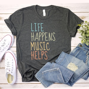 May include: Dark gray t-shirt with the text "LIFE HAPPENS MUSIC HELPS" in blue, green, orange, and pink. The shirt is paired with blue jeans and white sneakers with red accents. A sprig of green leaves is also visible.