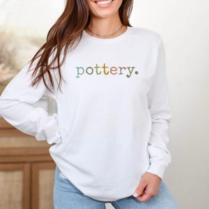 May include: A white long-sleeve shirt with the word "pottery." in a multi-colored font. The shirt is made of a soft material and is suitable for casual wear. The shirt is paired with blue jeans.