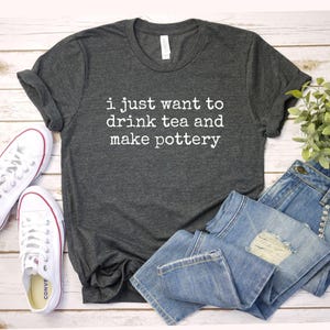 May include: Dark gray t-shirt with the text "i just want to drink tea and make pottery" in white. The shirt is paired with blue jeans and white sneakers with red accents, suggesting a casual, artistic theme.