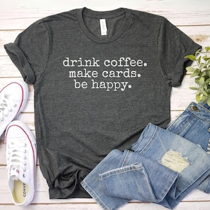 May include: A dark gray t-shirt with the text "drink coffee. make cards. be happy." printed in white.
