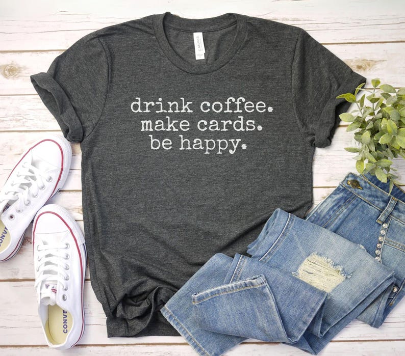 May include: A dark gray t-shirt with the text "drink coffee. make cards. be happy." printed in white.