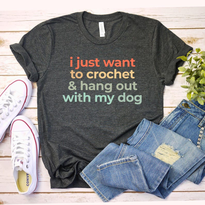 A gray t-shirt with text that says "I just want to crochet & hang out with my dog" is laid out on a wooden surface, accompanied by a pair of jeans and white sneakers.