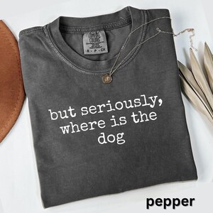 May include: A dark gray t-shirt with the phrase "but seriously, where is the dog" printed in white. The shirt has a crew neck and short sleeves. A silver necklace with a small pendant rests on the shirt. The word "pepper" is at the bottom.