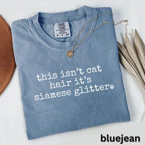 May include: Bluejean Comfort Colors t-shirt with the text "this isn't cat hair it's siamese glitter" in white. A gold necklace with a pendant rests on the shirt. The shirt has a small paw print graphic.