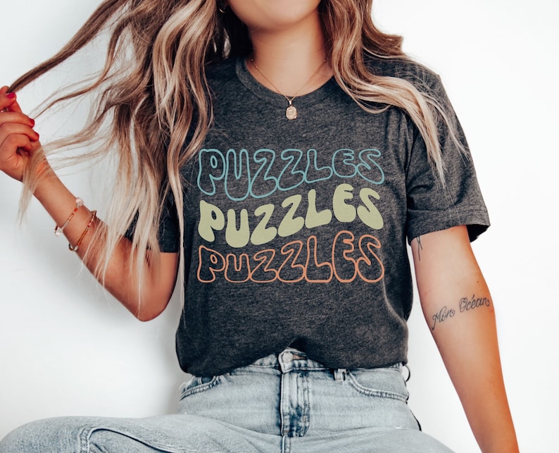 May include: A dark grey t-shirt with the word "PUZZLES" repeated three times in a retro font. Each word is a different colour: blue, green, and orange.