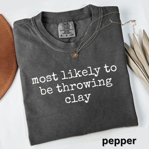 May include: A charcoal gray short-sleeve t-shirt with a crew neckline. The shirt features the white text "most likely to be throwing clay" printed on the front. A thin silver-toned chain necklace with a small circular pendant rests on the fabric.