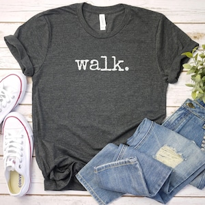 May include: A dark gray t-shirt with the word "walk." printed in white on the front. The shirt is laid flat on a white wooden surface with a pair of white sneakers and a pair of blue jeans.