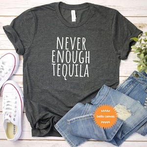May include: A dark gray t-shirt with the text "NEVER ENOUGH TEQUILA" in white lettering. The shirt is laid flat on a white wooden surface with a pair of white sneakers, blue jeans, and a green plant.