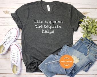 May include: A dark gray t-shirt with the text "life happens the tequila helps" printed in white on the front. The shirt is laid flat on a white wooden surface with a pair of white sneakers, blue jeans, and a small green plant.