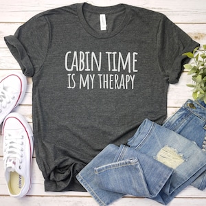 May include: A dark gray t-shirt with the text "Cabin Time Is My Therapy" in white lettering. The shirt is laid out on a white wooden surface with a pair of white sneakers and blue jeans.