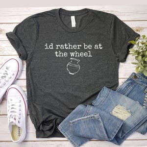 May include: Dark gray t-shirt with the text "id rather be at the wheel" and a white pottery wheel graphic. The shirt is paired with blue jeans and white canvas sneakers with red accents.