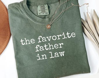 May include: A moss green t-shirt with the text "the favorite father in law" in white. A gold-colored necklace with a pendant is draped across the shirt. The shirt is folded on a white surface.