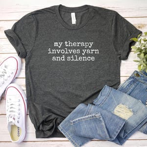 May include: A dark gray t-shirt with the text "my therapy involves yarn and silence" in white. The shirt is paired with light blue denim jeans and white canvas sneakers with red accents.