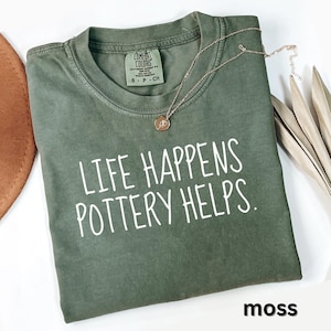May include: A moss green Comfort Colors t-shirt with the text "LIFE HAPPENS POTTERY HELPS" in white. A gold-colored necklace with a pendant rests on the shirt. The shirt is folded on a white surface.