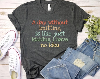 May include: A dark gray t-shirt with the text "A day without knitting is like, just kidding I have no idea" in rainbow colors. The shirt is laying on a white wooden surface with a pair of white sneakers and blue jeans.