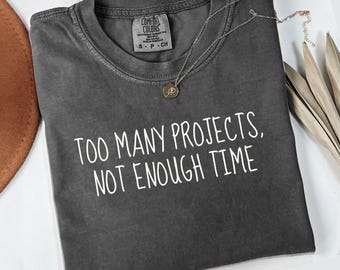 May include: A dark gray t-shirt with the text "TOO MANY PROJECTS, NOT ENOUGH TIME" in white. The shirt has a crew neck and a small tag on the upper left. A silver necklace with a pendant is draped across the shirt. The word "pepper" is at the bottom.