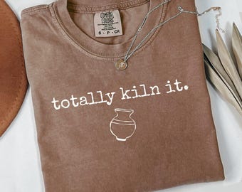 May include: Espresso-colored t-shirt with the text "totally kiln it." and a white illustration of a vase. A silver necklace with a pendant rests on the shirt. The shirt has a Comfort Colors label.
