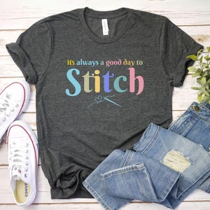 May include: A dark gray t-shirt with the text "it's always a good day to Stitch" in colorful letters. The text is accompanied by a small illustration of a needle and thread.