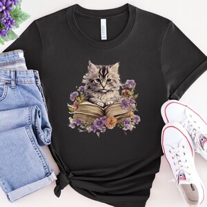 May include: A black t-shirt with a graphic of a gray and white kitten sitting on a book surrounded by purple, pink, and yellow flowers.