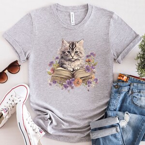 May include: Gray t-shirt with a graphic of a cat reading a book. The cat is gray and white with green eyes. The book is open and has a brown cover. There are flowers around the cat and book.