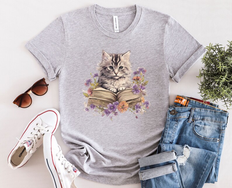 May include: Gray t-shirt with a graphic of a cat reading a book. The cat is gray and white with green eyes. The book is open and has a brown cover. There are flowers around the cat and book.