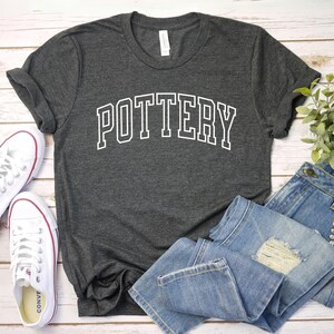 May include: A dark gray t-shirt with the word "POTTERY" printed in white on the front. The shirt is styled with blue jeans and white sneakers.