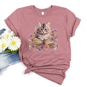 May include: A mauve t-shirt with a graphic of a cat sitting on an open book surrounded by flowers. The cat is gray and white with green eyes. The book is brown with a white page showing. The flowers are purple, pink, and yellow.