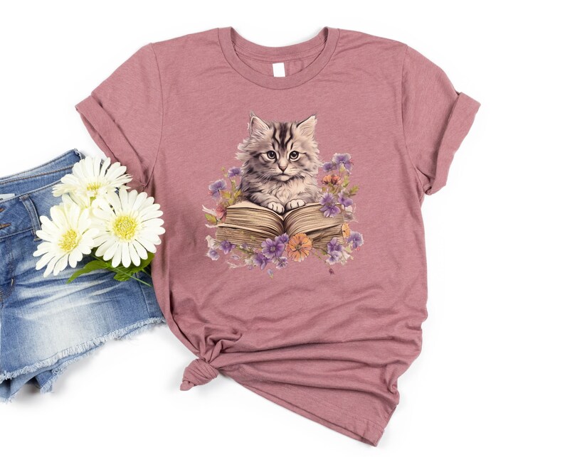 May include: A mauve t-shirt with a graphic of a cat sitting on an open book surrounded by flowers. The cat is gray and white with green eyes. The book is brown with a white page showing. The flowers are purple, pink, and yellow.