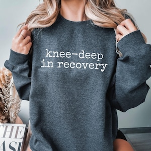 May include: Dark gray sweatshirt with the text "knee-deep in recovery" in white. The sweatshirt has long sleeves and a crew neck. The image shows a person wearing the sweatshirt.