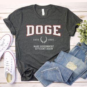 May include: A dark gray t-shirt with the text "DOGE" in white with a red outline. The text "ESTD 2025" is below the word "DOGE" with a laurel wreath between the two. The text "MAKE GOVERNMENT EFFICIENT AGAIN" is below the laurel wreath.
