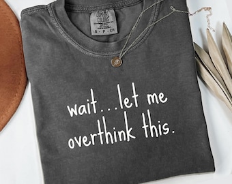 May include: A dark gray Comfort Colors t-shirt with the text "wait...let me overthink this" in white. A silver necklace with a pendant rests on the shirt. The word "pepper" is at the bottom.