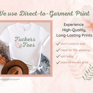 a white T-shirt with the text "Tuckers Tees" printed on it, along with a brown hat and some dried flowers. The image also includes text that describes the benefits of using Direct-to-Garment Print, such as a high-quality, long-lasting print that won't crack or peel, and a soft fabric state-of-the-art print.