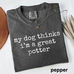 May include: A dark gray t-shirt with the text "my dog thinks I'm a great potter" in white. The shirt has a crew neck and a small tag on the upper left. A silver necklace with a pendant is on the shirt. The word "pepper" is at the bottom.